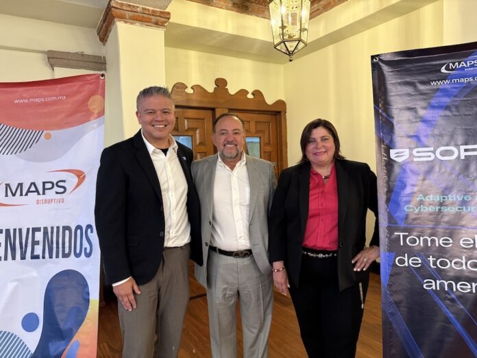 Alianza comercial estratégica entre MAPS y Sophos para impulsar la ciberseguridad unificada en México Fotografía en plano medio que muestra a tres directivos de MAPS y Sophos posando sonrientes de frente en un salón con pisos de madera y techo con vigas expuestas de ladrillo. A la izquierda, un ejecutivo de tez clara viste un saco oscuro sobre una camisa blanca y pantalón café, de pie junto a un banner publicitario vertical que muestra el logotipo de