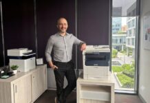Xerox integra a Lexmark para fortalecer el negocio del canal con un portafolio más amplio Fotografía en plano medio que muestra a Andrés Cid, product manager en Xerox, posando sonriente en una oficina corporativa. El ejecutivo viste una camisa de vestir gris claro de manga larga y pantalón negro. Se encuentra de pie, apoyando su brazo izquierdo sobre una impresora multifuncional blanca y azul de Xerox colocada sobre una mesa de madera clara. A la izquierda, sobre un archivador de oficina, se observa otro equipo de impresión de escritorio más pequeño y un organizador de plumas. De fondo, a la derecha, un gran ventanal ofrece vistas a un complejo de edificios corporativos modernos y áreas verdes.