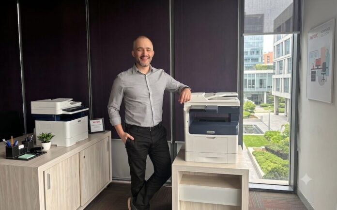 Fotografía en plano medio que muestra a Andrés Cid, product manager en Xerox, posando sonriente en una oficina corporativa. El ejecutivo viste una camisa de vestir gris claro de manga larga y pantalón negro. Se encuentra de pie, apoyando su brazo izquierdo sobre una impresora multifuncional blanca y azul de Xerox colocada sobre una mesa de madera clara. A la izquierda, sobre un archivador de oficina, se observa otro equipo de impresión de escritorio más pequeño y un organizador de plumas. De fondo, a la derecha, un gran ventanal ofrece vistas a un complejo de edificios corporativos modernos y áreas verdes.