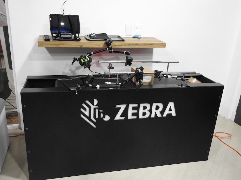 Fotografía que muestra un sistema modular de inspección por visión artificial montado sobre una estructura de soporte negra con la marca "ZEBRA" en letras blancas retroiluminadas al frente. Sobre la mesa metálica negra corren rieles horizontales cromados con brazos mecánicos articulados que sostienen cámaras de visión industrial fijas y sensores ópticos dirigidos hacia una caja de cartón pequeña. Al fondo, sobre una repisa flotante de madera empotrada en el muro blanco, se observa una tableta empresarial robusta colocada de forma vertical en su base de carga de escritorio.