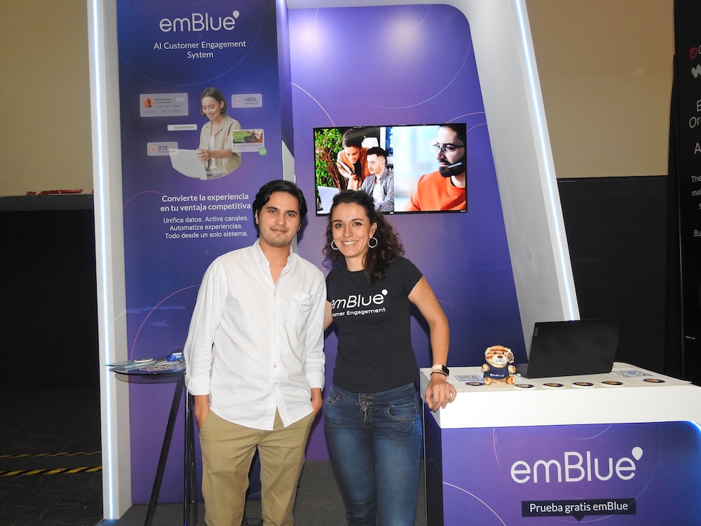 Fotografía de dos representantes de emBlue, un hombre con camisa blanca y una mujer con camiseta negra corporativa, posando sonrientes frente a un stand de exhibición morado. El fondo muestra el texto "emBlue AI Customer Engagement System" junto a los beneficios de unificar datos y activar canales. Sobre el mostrador blanco a su derecha hay una computadora portátil, posavasos y un pequeño tigre de peluche.