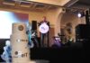 Grupo Be IT fortalece la ciberseguridad empresarial con un modelo integral que une IA, OT y talento especializado Fotografía en plano general medio con toma en ángulo contrapicado tomada desde un costado del escenario durante la ceremonia de aniversario. En primer plano a la izquierda resalta un gran pastel de bodas o de celebración de tres pisos color blanco con detalles decorativos en azul y el logotipo dorado de "Grupo Be IT" y "BURŌMC" en relieve. Al fondo, sobre el escenario oscuro iluminado por reflectores de teatro, el CEO Elías Cedillo lee un discurso oficial desde un podio transparente circular con el logotipo de la organización.