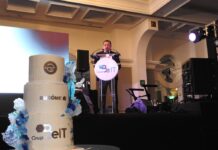 Grupo Be IT fortalece la ciberseguridad empresarial con un modelo integral que une IA, OT y talento especializado Fotografía en plano general medio con toma en ángulo contrapicado tomada desde un costado del escenario durante la ceremonia de aniversario. En primer plano a la izquierda resalta un gran pastel de bodas o de celebración de tres pisos color blanco con detalles decorativos en azul y el logotipo dorado de "Grupo Be IT" y "BURŌMC" en relieve. Al fondo, sobre el escenario oscuro iluminado por reflectores de teatro, el CEO Elías Cedillo lee un discurso oficial desde un podio transparente circular con el logotipo de la organización.