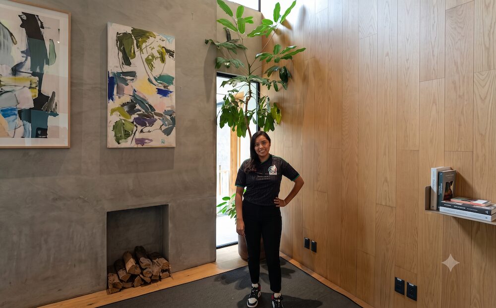 Fotografía de cuerpo completo en plano general que muestra a Citlali González, responsable de tesorería, administración y finanzas de Círculo Daat, posando de frente con las manos en los bolsillos de su pantalón. La ejecutiva viste una playera polo blanca oficial que tiene grabados los logotipos de "KANAN" y "Círculo Daat", pantalón negro y zapatos oscuros. Permanece de pie en el vestíbulo al lado de la chimenea minimalista empotrada en el muro de concreto gris, con el cuadro de arte abstracto azul y verde colgado arriba de esta.