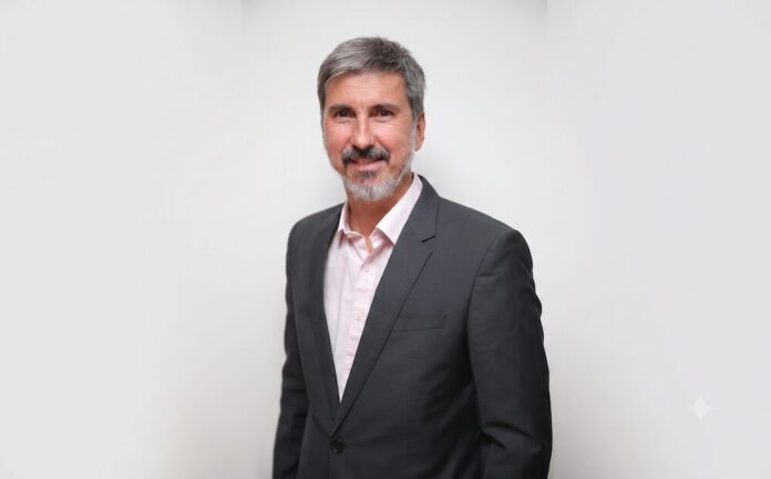Fotografía de medio cuerpo de Claudio Stopatto. Es un hombre de mediana edad con cabello entrecano y barba corta, que mira sonriente a la cámara. Viste un saco gris oscuro y una camisa de botones en tono rosa pálido, posando frente a un fondo blanco liso.