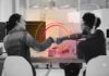 Splunk alerta que el costo del tiempo de inactividad ya alcanza 600 mil millones de dólares al año Fotografía en escala de grises que muestra a una mujer y a un hombre (usando audífonos) sentados frente a una computadora en un entorno de oficina. Ambos están chocando los puños sonrientes y, justo en el punto donde sus manos se encuentran, resalta un diseño gráfico superpuesto compuesto por aros concéntricos y líneas curvas brillantes en tonos magenta, naranja y rosa.