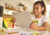 Sandisk y Crayola llevan la creatividad al almacenamiento con una colorida línea de memorias USB-C Fotografía de una niña (desenfocada en el fondo) sonriendo mientras sostiene una tableta plateada con ambas manos. Conectada al puerto lateral de la tableta destaca una unidad flash USB-C con forma de crayón color verde brillante (Electric Lime). Sobre la mesa, en primer plano, hay dibujos infantiles, crayones sueltos y una caja clásica de 64 crayones Crayola.