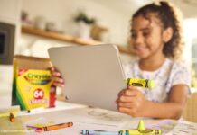Sandisk y Crayola llevan la creatividad al almacenamiento con una colorida línea de memorias USB-C Fotografía de una niña (desenfocada en el fondo) sonriendo mientras sostiene una tableta plateada con ambas manos. Conectada al puerto lateral de la tableta destaca una unidad flash USB-C con forma de crayón color verde brillante (Electric Lime). Sobre la mesa, en primer plano, hay dibujos infantiles, crayones sueltos y una caja clásica de 64 crayones Crayola.