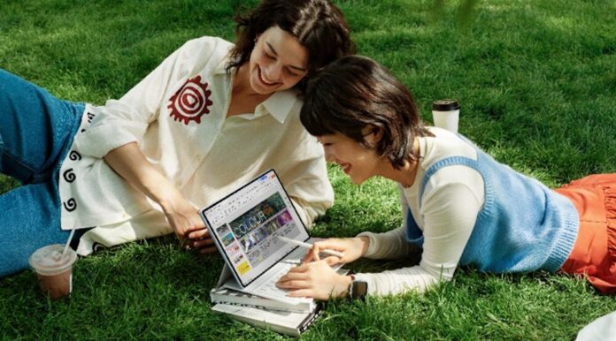 Dos mujeres jóvenes disfrutan de un día soleado recostadas sobre el césped de un parque. Una de ellas opera una tablet apoyada sobre unos libros, la cual está equipada con un teclado blanco y un lápiz óptico, mostrando un documento con imágenes coloridas en la pantalla. Ambas sonríen mientras miran el dispositivo.