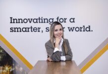 Axis Communications destaca cómo la videovigilancia evoluciona hacia una plataforma de inteligencia empresarial Fotografía de retrato de Diana Ardila, quien aparece sonriendo a la cámara con el rostro apoyado suavemente sobre su mano derecha, sentada frente a una mesa de madera. Viste una camisa blanca debajo de un saco color gris pardo. En el fondo, hay una pared blanca decorada con franjas diagonales en tonos ocre, marrón y gris, y se lee parcialmente el lema corporativo en letras negras: "Innovating for a smarter, s... world.".