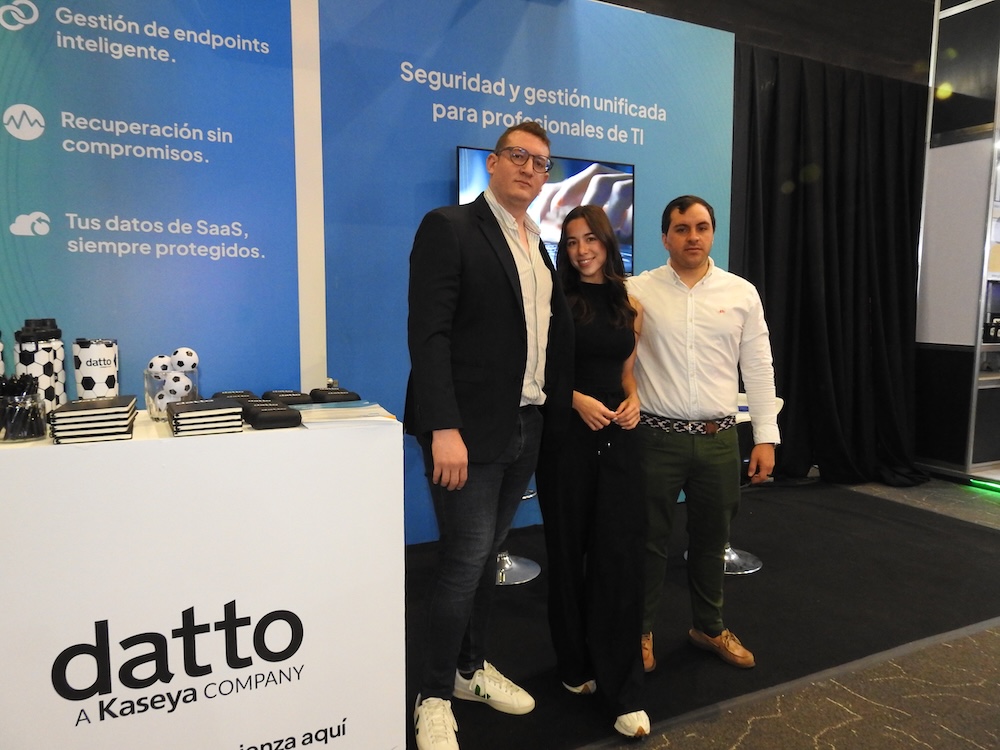 Plataforma de ciberseguridad para empresas:Fotografía en plano general medio de tres profesionales, dos hombres y una mujer, posando sonrientes de pie frente a un stand de exhibición. El mostrador blanco en primer plano muestra el logotipo negro de "datto A Kaseya COMPANY" junto con artículos promocionales como libretas y vasos con diseño de balón de fútbol. Al fondo, un panel azul resalta el texto "Seguridad y gestión unificada para profesionales de TI", acompañado de viñetas sobre protección de datos.