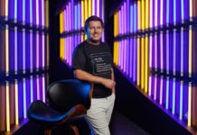 TOTVS destaca las lecciones que el Mundial dejó sobre tecnología, datos y gestión empresarial Fotografía de retrato de Javier Marbec, sonriendo a la cámara y apoyando su brazo derecho sobre el respaldo de una silla negra. Viste pantalones claros y una camiseta negra con un texto estampado que explica el significado de la marca "TOTVS". De fondo, se observa una instalación moderna con múltiples luces de neón verticales en colores morado, azul y amarillo.