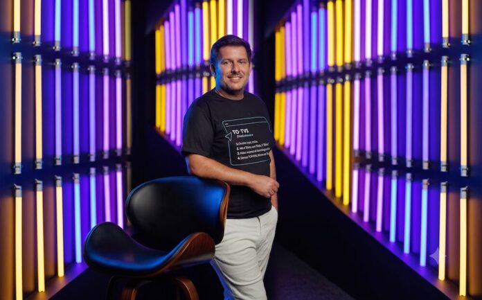 El legado del Mundial. Transformación digital y gestión integrada para el éxito corporativo Fotografía de retrato de Javier Marbec, sonriendo a la cámara y apoyando su brazo derecho sobre el respaldo de una silla negra. Viste pantalones claros y una camiseta negra con un texto estampado que explica el significado de la marca