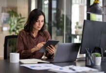 Flow impulsa la automatización inteligente para acelerar el crecimiento de las PyMEs mexicanas Fotografía de una mujer trabajando en una oficina moderna. Está sentada frente a su escritorio concentrada utilizando una tablet con ambas manos. Sobre la mesa de trabajo oscura se encuentran una laptop abierta, un monitor negro, una libreta de apuntes, una taza blanca y algunos documentos impresos con gráficas de negocios.