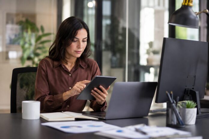 Fotografía de una mujer trabajando en una oficina moderna. Está sentada frente a su escritorio concentrada utilizando una tablet con ambas manos. Sobre la mesa de trabajo oscura se encuentran una laptop abierta, un monitor negro, una libreta de apuntes, una taza blanca y algunos documentos impresos con gráficas de negocios.