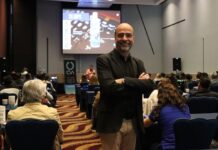 Grupo CVA fortalece al canal de TI con “CVA en tu Ciudad” 2026 en Puebla Fotografía de Zeferino Pérez, quien sonríe a la cámara de pie y con los brazos cruzados en el pasillo central de un salón de conferencias. Al fondo, los asistentes observan una presentación donde se proyecta la imagen de un producto de limpieza electrónica en la pantalla principal.