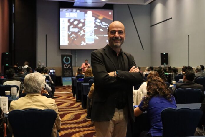 Fotografía de Zeferino Pérez, quien sonríe a la cámara de pie y con los brazos cruzados en el pasillo central de un salón de conferencias. Al fondo, los asistentes observan una presentación donde se proyecta la imagen de un producto de limpieza electrónica en la pantalla principal.