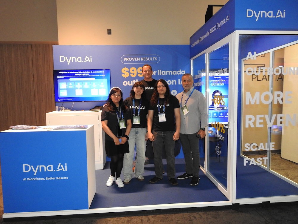 IA para contact center: Fotografía en plano general medio de cinco colaboradores de Dyna.Ai posando juntos de frente en su stand promocional durante una exposición tecnológica. El grupo está conformado por tres mujeres y dos hombres de perfiles intergeneracionales; cuatro de ellos visten playeras tipo polo negras corporativas con el logotipo bordado de "Dyna.Ai" y portan gafetes del evento colgados del cuello, mientras que el hombre del extremo derecho viste una camisa gris claro de vestir. Al fondo, el stand azul rey cuenta con un mostrador blanco, una pantalla digital con diagramas de flujos de trabajo agénticos y un letrero que dice "Centro de Demo de AICC Dyna.Ai", flanqueado por una cabina de cristal con la leyenda caligráfica "MORE REVENUE SCALE FAST".