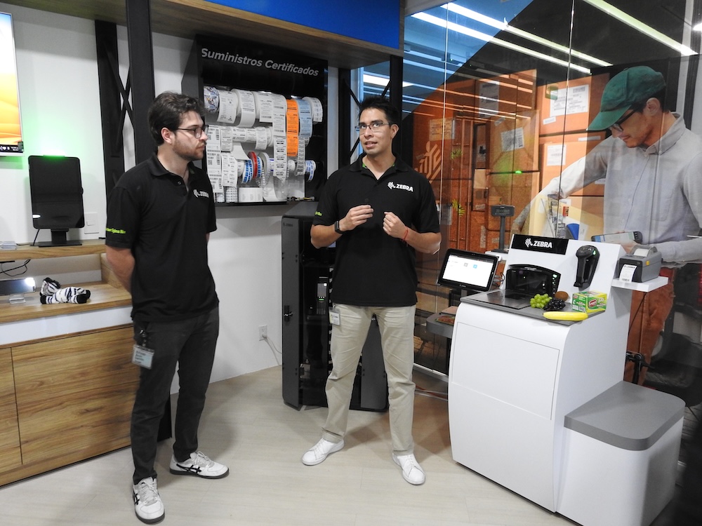 Fotografía en plano general medio que muestra a dos ingenieros de Zebra Technologies de pie ofreciendo una demostración en un espacio interactivo de showroom. A la izquierda, un técnico joven con lentes y playera tipo polo negra de Zebra observa la estación de trabajo. Al centro, el segundo ingeniero, vestido igualmente con la playera corporativa negra, pantalón beige y tenis blancos, gesticula con las manos mientras explica el sistema. A la derecha se sitúa un mostrador de punto de venta blanco que integra un escáner de base con frutas artificiales (plátano y uvas) sobre la báscula, una pantalla táctil, una terminal móvil de captura y una pequeña impresora de etiquetas, teniendo como fondo una mampara con suministros certificados y un gráfico de un operador logístico.