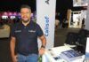 Calisof impulsa una nueva etapa en la gestión inteligente de TI con ITSM Fotografía de medio cuerpo de Ericcsson Pinto, parado frente a un mostrador blanco en un stand de exposición corporativa. Viste una playera tipo polo azul marino con los logotipos de Acronis y Calisof bordados en el pecho, junto con pantalones de mezclilla azul claro. Su mano derecha descansa en su bolsillo mientras la izquierda se apoya sobre el mostrador, donde hay apilados folletos informativos. Al fondo, se observa el entorno iluminado de una feria tecnológica con otras personas y stands.