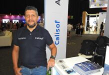 Calisof ITSM impulsa una nueva etapa para la gestión inteligente de TI Fotografía de medio cuerpo de Ericcsson Pinto, parado frente a un mostrador blanco en un stand de exposición corporativa. Viste una playera tipo polo azul marino con los logotipos de Acronis y Calisof bordados en el pecho, junto con pantalones de mezclilla azul claro. Su mano derecha descansa en su bolsillo mientras la izquierda se apoya sobre el mostrador, donde hay apilados folletos informativos. Al fondo, se observa el entorno iluminado de una feria tecnológica con otras personas y stands.