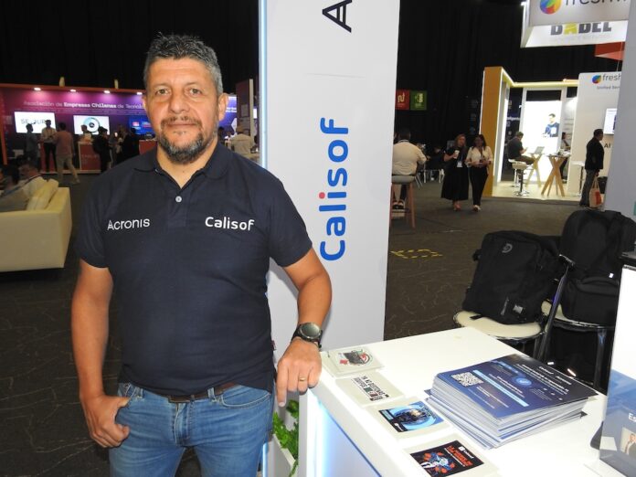 Fotografía de medio cuerpo de Ericcsson Pinto, parado frente a un mostrador blanco en un stand de exposición corporativa. Viste una playera tipo polo azul marino con los logotipos de Acronis y Calisof bordados en el pecho, junto con pantalones de mezclilla azul claro. Su mano derecha descansa en su bolsillo mientras la izquierda se apoya sobre el mostrador, donde hay apilados folletos informativos. Al fondo, se observa el entorno iluminado de una feria tecnológica con otras personas y stands.