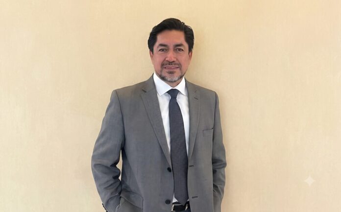 Retrato de estudio en plano medio y de frente de Fausto Escobar, CEO de HD Latinoamérica, posando de pie con una sutil sonrisa y las manos en los bolsillos de su pantalón. El ejecutivo, de tez morena clara, cabello oscuro y barba corta de candado bien recortada, viste un saco de traje formal color gris, una camisa de vestir blanca de cuello rígido y una corbata de color gris oscuro texturizada, situado ante un muro liso de fondo beige claro.