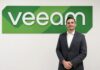 Fernando Zambrana liderará la estrategia de Veeam México para impulsar la resiliencia de datos Retrato corporativo de plano medio y de frente de Fernando Zambrana, Country Director de Veeam en México, posando sonriente ante un muro liso de fondo blanco. El ejecutivo, de tez clara y cabello corto oscuro, viste un saco formal gris jaspeado, una camisa de vestir blanca de cuello rígido desabrochada en el primer botón y mantiene las manos cruzadas al frente. Detrás de él, en la sección superior izquierda, resalta un gran letrero institucional tridimensional con la palabra "veeam" en letras blancas de relieve, montadas sobre una base geométrica alargada con franjas diagonales en distintas tonalidades de verde brillante.