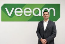 Fernando Zambrana liderará la estrategia de Veeam México para impulsar la resiliencia de datos Retrato corporativo de plano medio y de frente de Fernando Zambrana, Country Director de Veeam en México, posando sonriente ante un muro liso de fondo blanco. El ejecutivo, de tez clara y cabello corto oscuro, viste un saco formal gris jaspeado, una camisa de vestir blanca de cuello rígido desabrochada en el primer botón y mantiene las manos cruzadas al frente. Detrás de él, en la sección superior izquierda, resalta un gran letrero institucional tridimensional con la palabra "veeam" en letras blancas de relieve, montadas sobre una base geométrica alargada con franjas diagonales en distintas tonalidades de verde brillante.