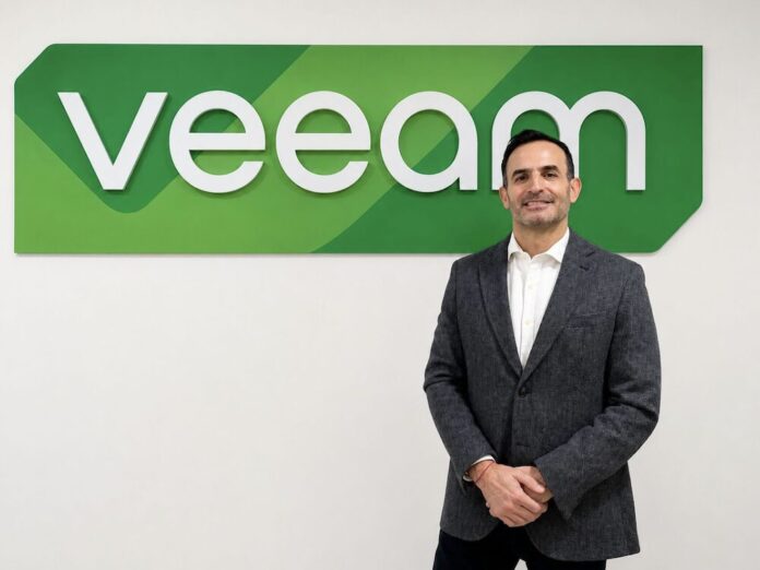 Fernando Zambrana asume la dirección de Veeam en México para potenciar la resiliencia de datos corporativos. Retrato corporativo de plano medio y de frente de Fernando Zambrana, Country Director de Veeam en México, posando sonriente ante un muro liso de fondo blanco. El ejecutivo, de tez clara y cabello corto oscuro, viste un saco formal gris jaspeado, una camisa de vestir blanca de cuello rígido desabrochada en el primer botón y mantiene las manos cruzadas al frente. Detrás de él, en la sección superior izquierda, resalta un gran letrero institucional tridimensional con la palabra