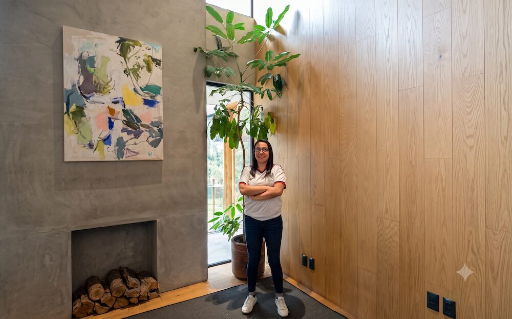 Ciberseguridad OT: Fotografía de cuerpo completo en plano general que muestra a Gabriela Almonte, coordinadora de capital humano de Círculo Daat, posando de frente con los brazos cruzados en el vestíbulo de diseño contemporáneo. La especialista tiene cabello largo oscuro, usa anteojos y viste una playera deportiva blanca de cuello tipo polo con detalles negros y rojos, jeans de mezclilla azul oscuro ajustados y tenis blancos. Se encuentra parada en el centro del espacio, frente a un ventanal alto de fondo que da hacia un patio interior, teniendo la pared de concreto gris a la izquierda y el muro de paneles de madera clara a la derecha.