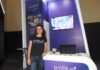 emBlue impulsa el customer engagement con datos centralizados y automatización inteligente Retrato de cuerpo entero de Giulia Landis, de pie junto al mostrador del stand de emBlue. Viste pantalones de mezclilla y una camiseta negra con la leyenda "emBlue Customer Engagement". A su espalda, una pantalla empotrada en la pared morada del stand proyecta una imagen con los iconos digitales de Email, SMS y WhatsApp.