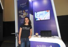 emBlue impulsa el customer engagement con datos centralizados y automatización inteligente Retrato de cuerpo entero de Giulia Landis, de pie junto al mostrador del stand de emBlue. Viste pantalones de mezclilla y una camiseta negra con la leyenda "emBlue Customer Engagement". A su espalda, una pantalla empotrada en la pared morada del stand proyecta una imagen con los iconos digitales de Email, SMS y WhatsApp.