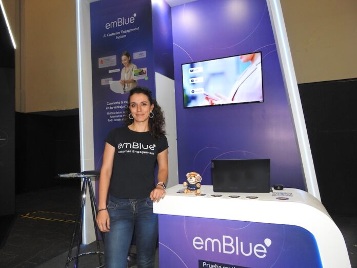 Giulia Landis detalla la integración omnicanal para el customer engagement Retrato de cuerpo entero de Giulia Landis, de pie junto al mostrador del stand de emBlue. Viste pantalones de mezclilla y una camiseta negra con la leyenda