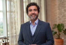 Fortinet: La ola llegó antes de lo previsto (y no la trajo un humano) Fotografía de medio cuerpo de Gonzalo Garcia, un hombre sonriente con cabello oscuro rizado y barba corta. Viste un traje gris oscuro, chaleco a juego y camisa celeste sin corbata. El fondo muestra una oficina moderna e iluminada, con un gran ventanal a la izquierda que deja ver las copas de los árboles, y una planta decorativa de hojas grandes a la derecha.