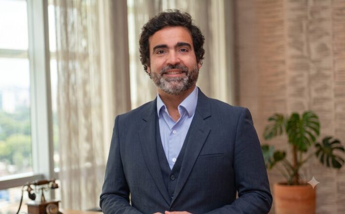 Fotografía de medio cuerpo de Gonzalo Garcia, un hombre sonriente con cabello oscuro rizado y barba corta. Viste un traje gris oscuro, chaleco a juego y camisa celeste sin corbata. El fondo muestra una oficina moderna e iluminada, con un gran ventanal a la izquierda que deja ver las copas de los árboles, y una planta decorativa de hojas grandes a la derecha.
