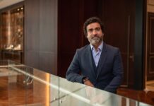 Fortinet anticipa que la próxima ola en ciberseguridad no será de ataques, sino de parches Retrato corporativo de plano medio y de frente de Gonzalo García, Vicepresidente de Fortinet para Sudamérica, posando sonriente en un entorno de oficina de diseño contemporáneo. El ejecutivo tiene cabello oscuro y una barba corta y canosa bien cuidada; viste un traje formal de tres piezas en color azul marino texturizado, con saco de dos botones, chaleco y camisa azul claro de cuello abierto, sin corbata. García permanece de pie, apoyando su brazo izquierdo sobre un barandal de cristal transparente, mientras que en la muñeca derecha se aprecia un reloj inteligente. De fondo, de manera difuminada, se observa un pasillo con pisos de madera, muros revestidos en madera oscura, una puerta elegante y una exhibición iluminada al fondo.