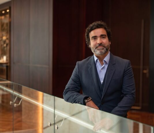 Fortinet anticipa que la próxima ola en ciberseguridad no será de ataques, sino de parches Retrato corporativo de plano medio y de frente de Gonzalo García, Vicepresidente de Fortinet para Sudamérica, posando sonriente en un entorno de oficina de diseño contemporáneo. El ejecutivo tiene cabello oscuro y una barba corta y canosa bien cuidada; viste un traje formal de tres piezas en color azul marino texturizado, con saco de dos botones, chaleco y camisa azul claro de cuello abierto, sin corbata. García permanece de pie, apoyando su brazo izquierdo sobre un barandal de cristal transparente, mientras que en la muñeca derecha se aprecia un reloj inteligente. De fondo, de manera difuminada, se observa un pasillo con pisos de madera, muros revestidos en madera oscura, una puerta elegante y una exhibición iluminada al fondo.