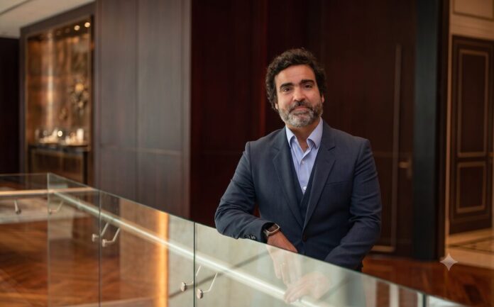 Retrato corporativo de plano medio y de frente de Gonzalo García, Vicepresidente de Fortinet para Sudamérica, posando sonriente en un entorno de oficina de diseño contemporáneo. El ejecutivo tiene cabello oscuro y una barba corta y canosa bien cuidada; viste un traje formal de tres piezas en color azul marino texturizado, con saco de dos botones, chaleco y camisa azul claro de cuello abierto, sin corbata. García permanece de pie, apoyando su brazo izquierdo sobre un barandal de cristal transparente, mientras que en la muñeca derecha se aprecia un reloj inteligente. De fondo, de manera difuminada, se observa un pasillo con pisos de madera, muros revestidos en madera oscura, una puerta elegante y una exhibición iluminada al fondo.
