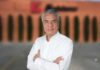 Inversión manufacturera de Koblenz fortalece su apuesta industrial con una expansión de largo alcance Retrato corporativo de plano medio y de frente de Héctor Lara, director de Marketing de Koblenz, posando con los brazos cruzados. El ejecutivo, de tez clara y cabello corto canoso peinado hacia atrás, viste una camisa formal blanca de manga larga. Al fondo, de manera difuminada y fuera de foco, se observa la fachada de ladrillo de una nave industrial con árboles esbeltos alineados y un logotipo corporativo rojizo parcialmente visible.