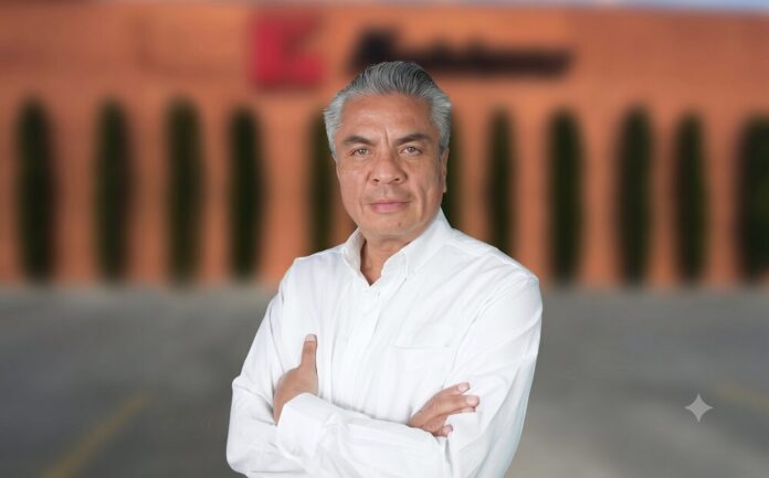 Retrato corporativo de plano medio y de frente de Héctor Lara, director de Marketing de Koblenz, posando con los brazos cruzados. El ejecutivo, de tez clara y cabello corto canoso peinado hacia atrás, viste una camisa formal blanca de manga larga. Al fondo, de manera difuminada y fuera de foco, se observa la fachada de ladrillo de una nave industrial con árboles esbeltos alineados y un logotipo corporativo rojizo parcialmente visible.