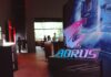 Gigabyte celebra 40 años con una nueva estrategia para fortalecer al canal Fotografía en plano detalle con enfoque selectivo del letrero acrílico de AORUS iluminado en colores neón azul y morado, montado sobre una base negra mate texturizada. El fondo de la toma muestra un salón de eventos corporativo difuminado de forma artística, donde se aprecian las siluetas de asistentes conversando y mamparas publicitarias con luces rojas y blancas.
