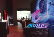 Gigabyte celebra 40 años con una nueva estrategia para fortalecer al canal Fotografía en plano detalle con enfoque selectivo del letrero acrílico de AORUS iluminado en colores neón azul y morado, montado sobre una base negra mate texturizada. El fondo de la toma muestra un salón de eventos corporativo difuminado de forma artística, donde se aprecian las siluetas de asistentes conversando y mamparas publicitarias con luces rojas y blancas.