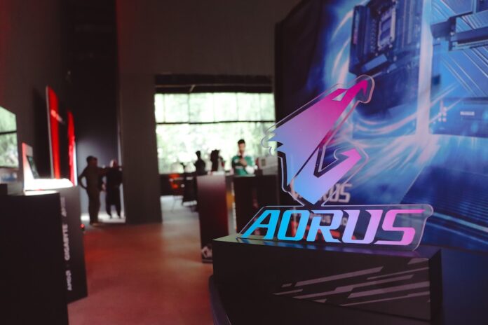 Fotografía en plano detalle con enfoque selectivo del letrero acrílico de AORUS iluminado en colores neón azul y morado, montado sobre una base negra mate texturizada. El fondo de la toma muestra un salón de eventos corporativo difuminado de forma artística, donde se aprecian las siluetas de asistentes conversando y mamparas publicitarias con luces rojas y blancas.