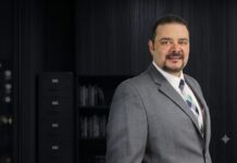 Perfect Choice fortalece sus canales de distribución con una estrategia basada en cercanía, innovación y crecimiento Retrato de medio cuerpo de Armando Gallo posando en una oficina con iluminación tenue. Lleva un traje gris, camisa blanca y corbata con patrón de cuadros en tonos azules y verdes. El fondo oscuro muestra de manera difuminada un archivero y un librero negro.