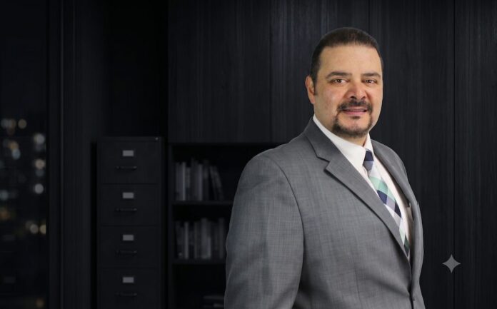 Retrato de medio cuerpo de Armando Gallo posando en una oficina con iluminación tenue. Lleva un traje gris, camisa blanca y corbata con patrón de cuadros en tonos azules y verdes. El fondo oscuro muestra de manera difuminada un archivero y un librero negro.