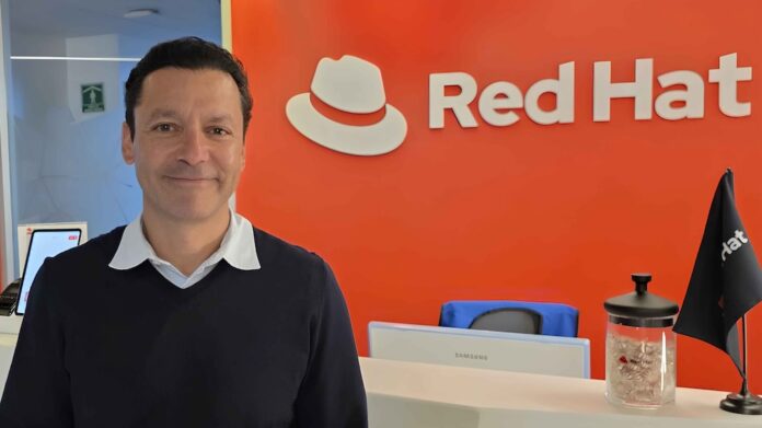 Jorge Pastrana asume el liderazgo del segmento comercial de Red Hat para América Latina Fotografía en plano medio de Jorge Pastrana, quien posa sonriendo a la cámara de forma relajada. Viste un suéter oscuro con cuello en V sobre una camisa blanca. De fondo, se observa la recepción de una oficina corporativa protagonizada por una gran pared de color naranja vibrante que exhibe el icónico logotipo del sombrero blanco y el nombre