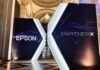 Epson apuesta por México para acelerar el crecimiento de su negocio corporativo y fortalecer a sus canales Estructura publicitaria iluminada con forma de letra "X" gigante, ubicada en el pasillo de un recinto de estilo histórico. El panel izquierdo muestra la palabra "EPSON" y el derecho "PARTNERX", ambos impresos sobre un fondo azul oscuro con un diseño gráfico de nodos digitales.