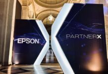 Epson apuesta por México para acelerar el crecimiento de su negocio corporativo y fortalecer a sus canales Estructura publicitaria iluminada con forma de letra "X" gigante, ubicada en el pasillo de un recinto de estilo histórico. El panel izquierdo muestra la palabra "EPSON" y el derecho "PARTNERX", ambos impresos sobre un fondo azul oscuro con un diseño gráfico de nodos digitales.