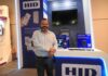 HID impulsa las identidades seguras con un ecosistema que transforma el control de acceso Fotografía de un hombre sonriente, Luis Yáñez, con camisa celeste y pantalón oscuro, de pie frente a un stand de exhibición de la marca HID. El módulo, iluminado con luces LED azules, muestra letreros promocionales sobre alianzas tecnológicas y exhibe dispositivos de seguridad como un lector de reconocimiento facial (HID Amico) y un lector de acceso móvil (HID Mobile Access) sobre un mostrador blanco.